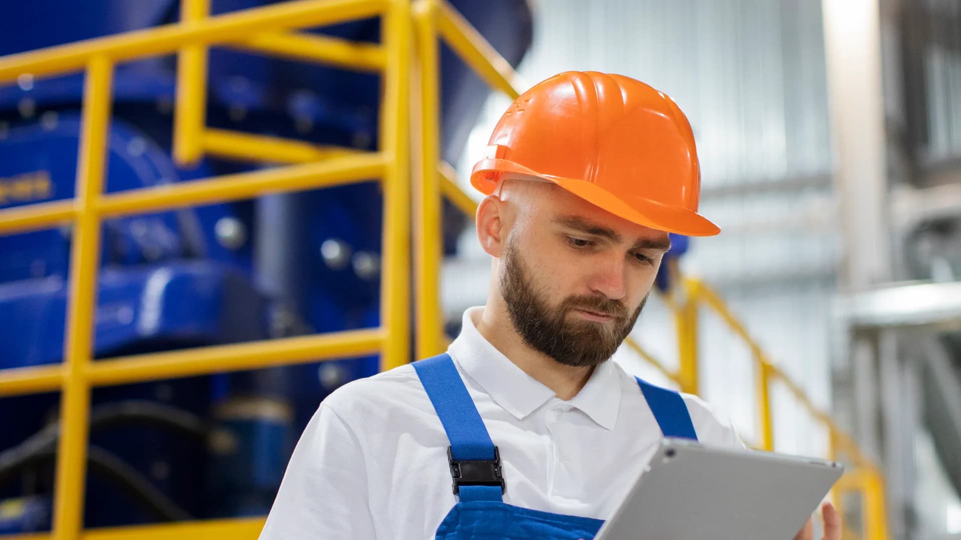 Operario con casco naranja y peto azul consulta una tablet en una planta industrial, en lugar de un parte de trabajo en papel